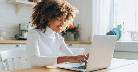 A woman wearing a white turtleneck sweater sits at a wooden kitchen table, looking at the screen of an open laptop.