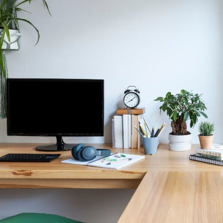 An L-shaped wooden desk features a desktop computer monitor, small potted plants, headphones, books, and a cellphone.