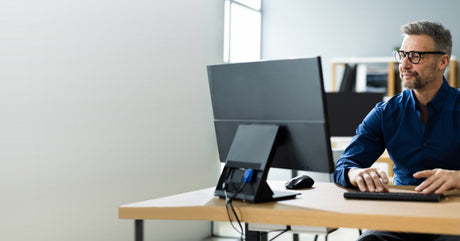 A happy man sits at a wooden desk typing on a black wireless keyboard and looking at the desktop monitor.