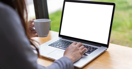 A close-up view shows a person sitting at a table holding a white mug in one hand and touching a laptop keyboard with the other.